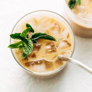 Clear glass with ice cubes, creamy vanilla coffee, and a fresh bundle of mint floating atop next to a silver spoon.